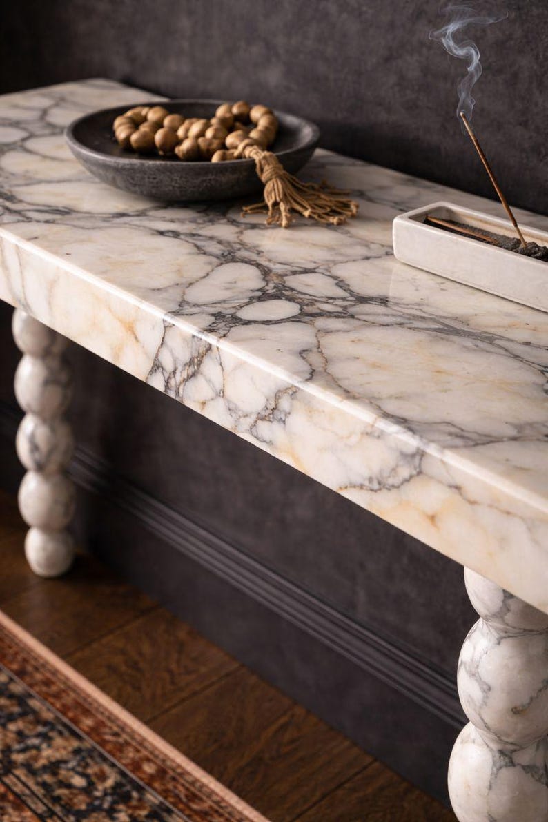 May include: A marble-topped console table with a grey and cream veined pattern. The table has turned legs and is decorated with a dark grey bowl holding prayer beads and a white incense holder. Incense smoke rises from the holder.
