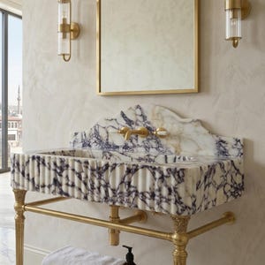 Pu&ograve; includere: Un lavabo in marmo a parete con venature bianche e viola e un design frontale scanalato. Il lavabo &egrave; sostenuto da una struttura in metallo dorato con gambe decorative e include un ripiano in marmo abbinato con un asciugamano bianco.
