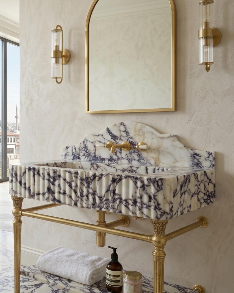 Pu&ograve; includere: Un lavabo in marmo a parete con venature bianche e viola e un design frontale scanalato. Il lavabo &egrave; sostenuto da una struttura in metallo dorato con gambe decorative e include un ripiano in marmo abbinato con un asciugamano bianco.