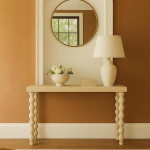 May include: A beige console table with a lamp, a bowl of flowers, and a small box. A round mirror hangs above the table, set against a warm brown wall. The table has decorative, rounded legs, and the room has a neutral colour palette.