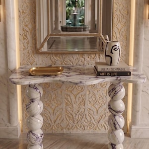 May include: A white marble console table with purple veining, featuring a wavy top and two legs composed of stacked spheres. On the table sits a gold rectangular tray and a white vase with black line art next to a book titled Julian Schnabel Art Is Life.