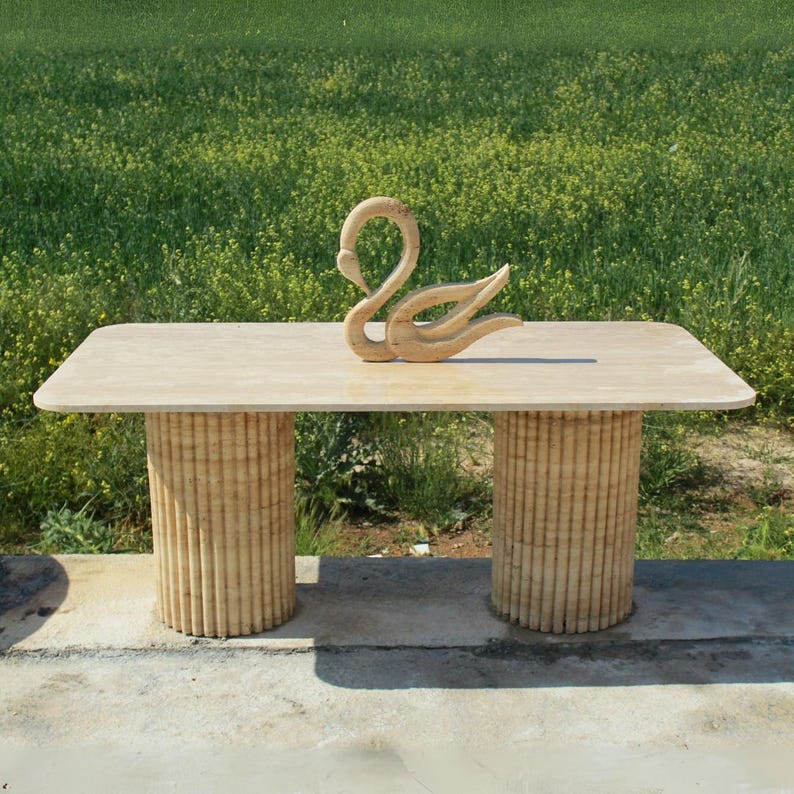 Puede incluir: Mesa rectangular beige con dos bases cil&iacute;ndricas. Una escultura de cisne de madera tallada se encuentra sobre la mesa. La mesa es de material claro, con un fondo de campo verde.