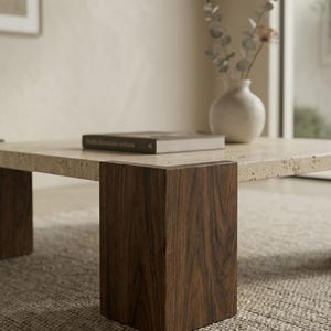 May include: A square coffee table featuring a porous beige travertine stone top supported by four thick, dark brown walnut wood legs. A single rectangular book with a dark cover rests on the stone surface next to a white ceramic vase.