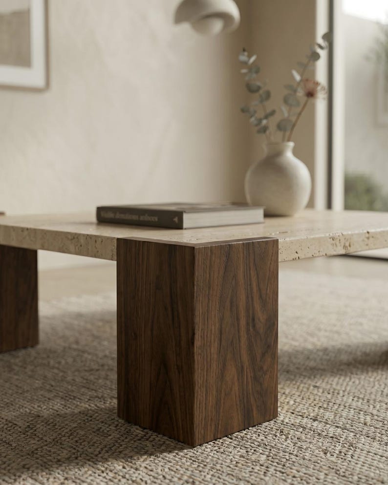 May include: A square coffee table featuring a porous beige travertine stone top supported by four thick, dark brown walnut wood legs. A single rectangular book with a dark cover rests on the stone surface next to a white ceramic vase.