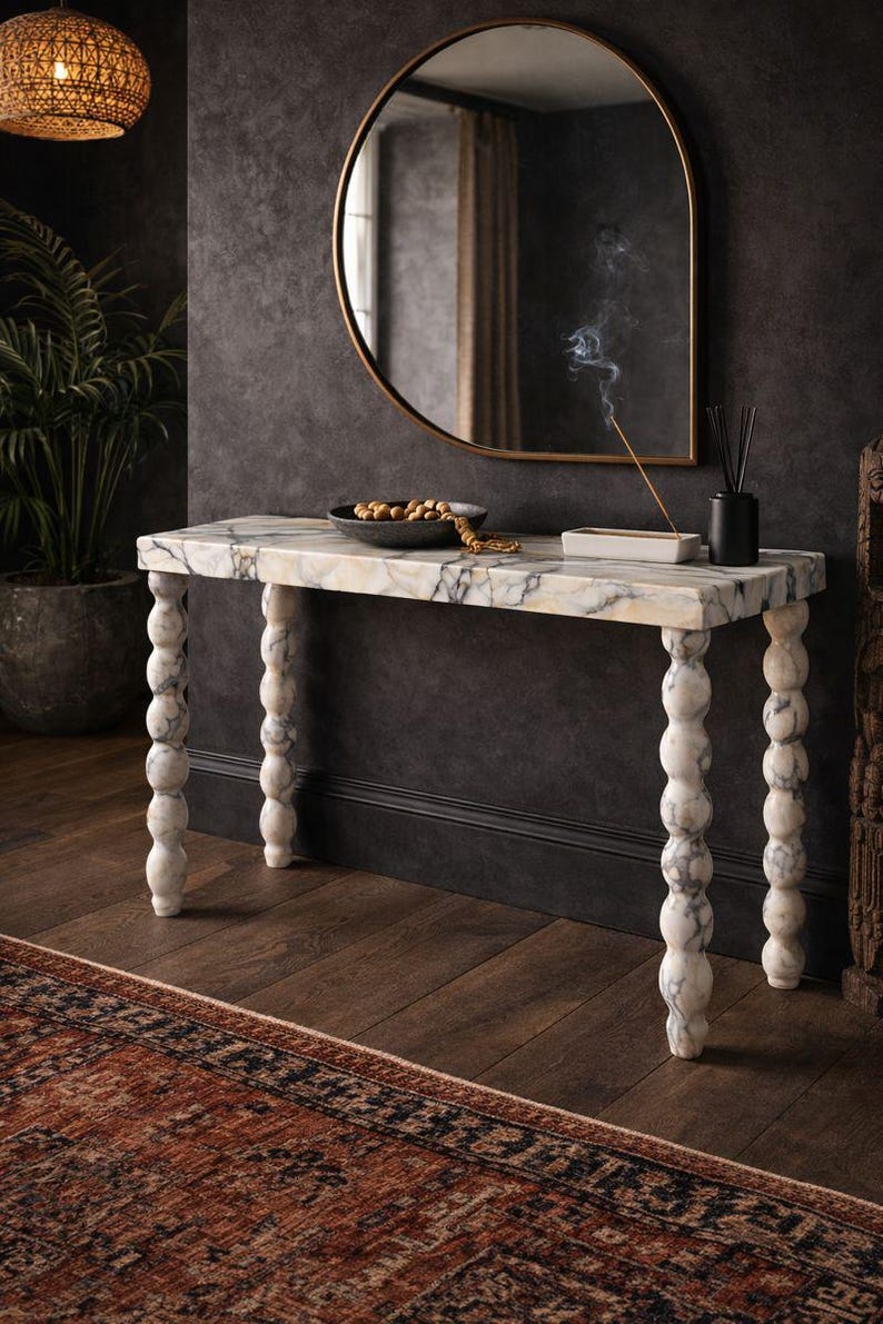 May include: A marble-topped console table with turned, stacked-ball legs. The table has a white and grey veined pattern. A round gold-framed mirror hangs above the table. A decorative bowl and incense burner sit on the table.