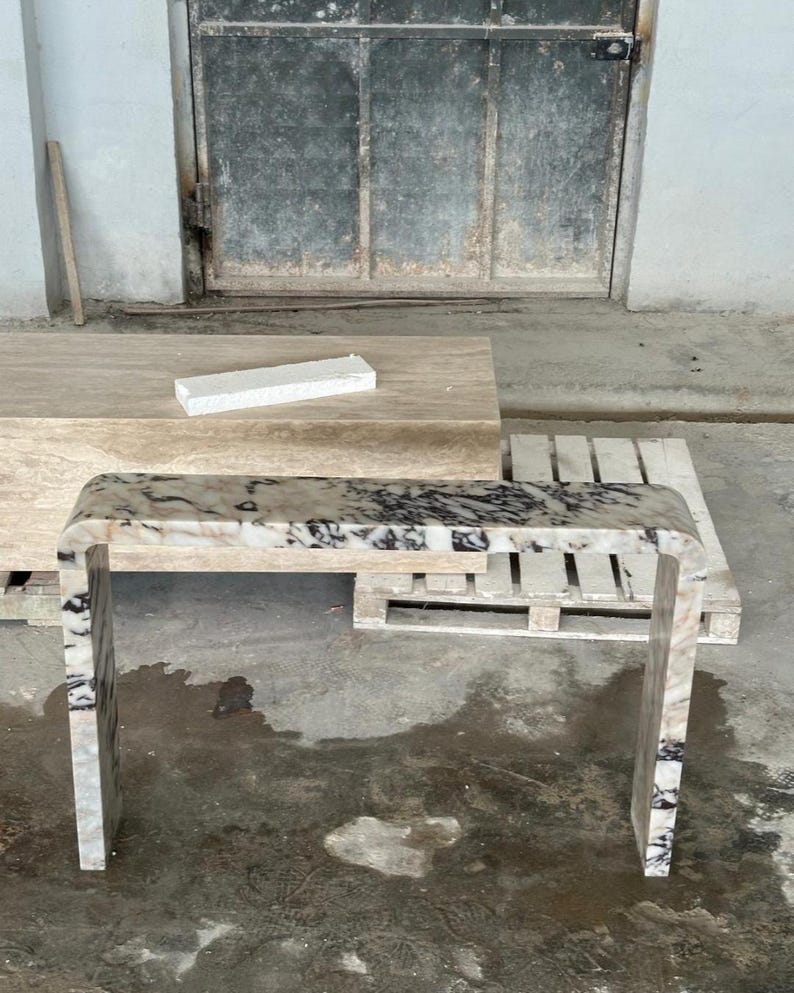 May include: A waterfall console table crafted from polished stone with a distinct white, beige, and black marbled pattern. The table features a continuous, smooth U-shaped design with rounded edges, showcasing natural veining throughout the surface.