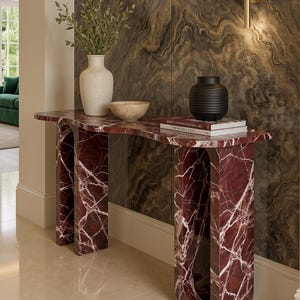 Op de afbeelding: Een dieprode marmeren sidetable met witte aders, voorzien van twee boogvormige poten met open centra. Op het oppervlak staan een hoge gebroken witte vaas met groen, een kleine beige kom, een zwarte geribbelde vaas en twee op elkaar gestapelde boeken.