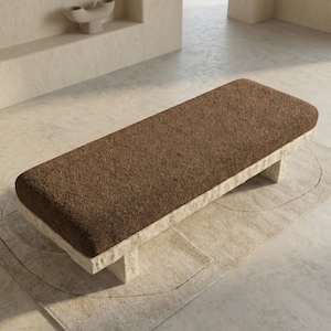 May include: A rectangular bench featuring a thick, dark brown textured fabric seat cushion supported by a sturdy, light beige stone base with a rough, natural finish. The bench is positioned horizontally, showcasing a contrast between the soft top and the solid, rugged stone structure underneath.