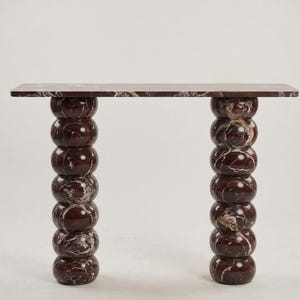May include: A modern console table with a rectangular top and two stacked, spherical legs. The table is made of a dark red marble with white veining. The table's dimensions are not visible.