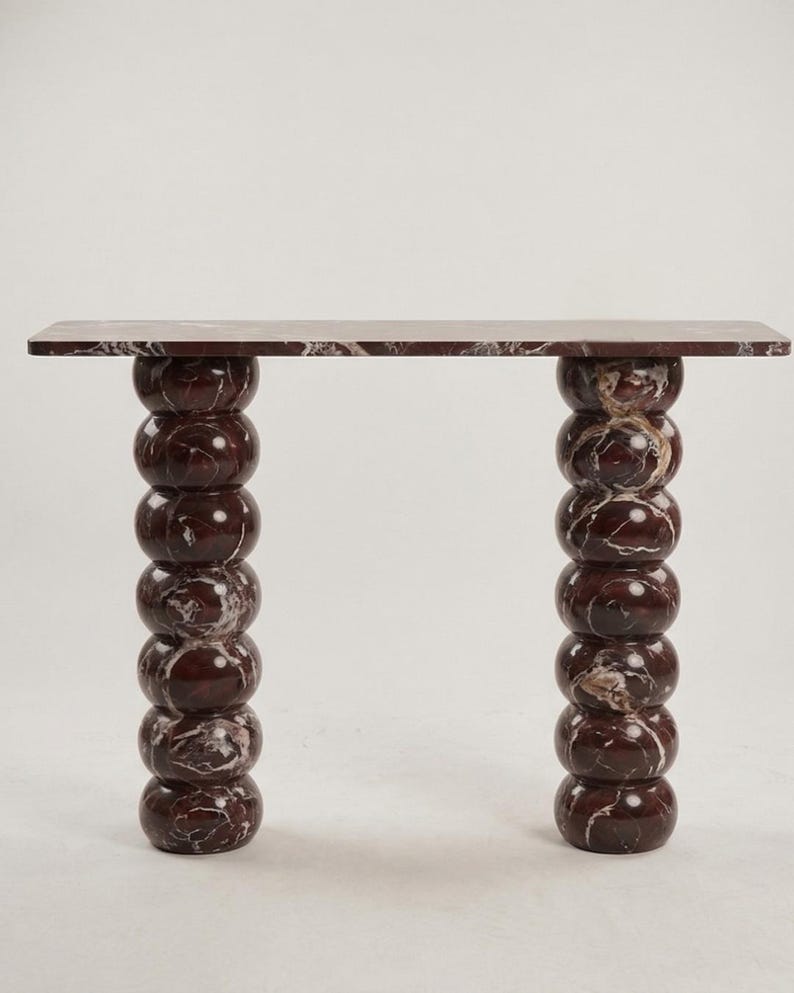 May include: A modern console table with a rectangular top and two stacked, spherical legs. The table is made of a dark red marble with white veining. The table's dimensions are not visible.