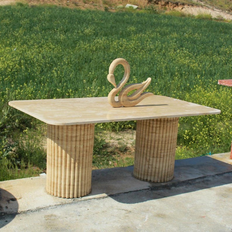 Puede incluir: Una mesa rectangular beige con dos patas cil&iacute;ndricas y acanaladas. Una escultura de cisne de madera tallada se encuentra sobre la superficie de la mesa. La mesa est&aacute; al aire libre, con un campo verde al fondo.