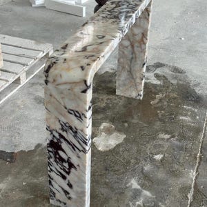 May include: A waterfall console table crafted from polished marble featuring a cream base color with prominent dark brown and black veining throughout the surface. The piece has a smooth, continuous curved edge connecting the top to the side leg.