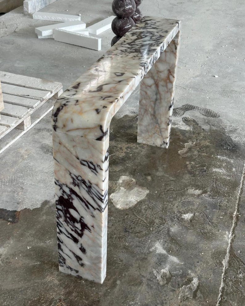 May include: A waterfall console table crafted from polished marble featuring a cream base color with prominent dark brown and black veining throughout the surface. The piece has a smooth, continuous curved edge connecting the top to the side leg.