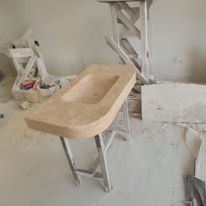 May include: A light beige rectangular bathroom sink with a rounded front edge, set on a white metal stand. The sink has a rectangular basin and two faucet holes. The background shows construction materials and tools.