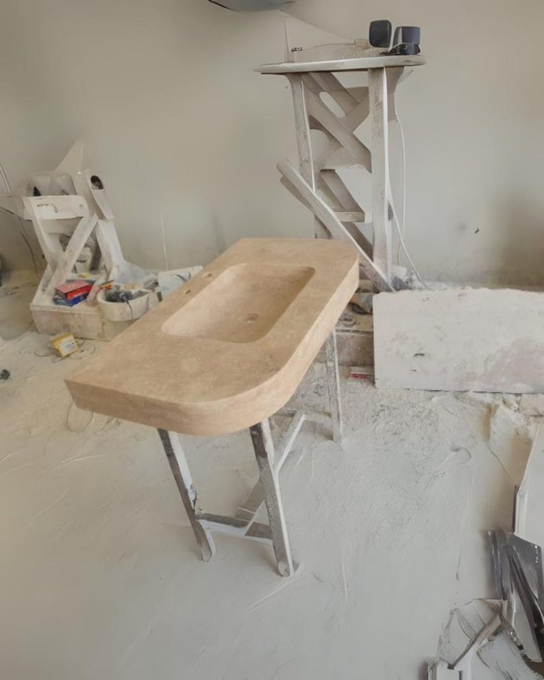 May include: A light beige rectangular bathroom sink with a rounded front edge, set on a white metal stand. The sink has a rectangular basin and two faucet holes. The background shows construction materials and tools.