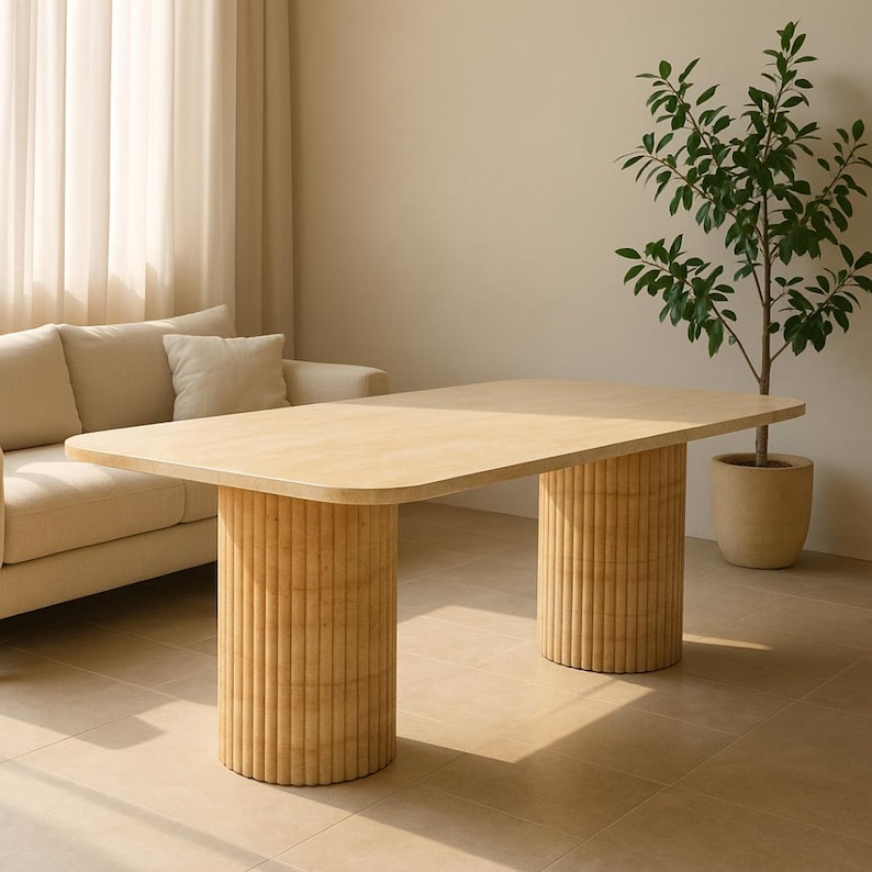 Puede incluir: Una mesa de comedor de madera clara con esquinas redondeadas y dos columnas de soporte cil&iacute;ndricas estriadas. La mesa mide aproximadamente 183 cm de largo. Un sof&aacute; beige y una planta en maceta est&aacute;n en el fondo.