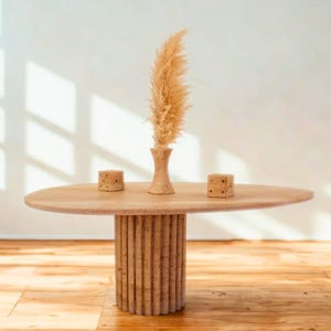 Puede incluir: Una mesa de centro de madera clara con una tapa ovalada y una base cilíndrica. La mesa presenta un pequeño jarrón con hierba ornamental seca y dos pequeños dados. La mesa está sobre un suelo de madera.