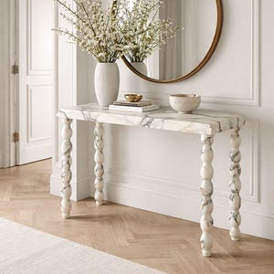 May include: A marble-topped console table with turned legs, a round mirror, and a vase of white flowers. The table has a rectangular top with grey veining and decorative, white and grey legs. A gold bowl and a textured bowl sit on the table.