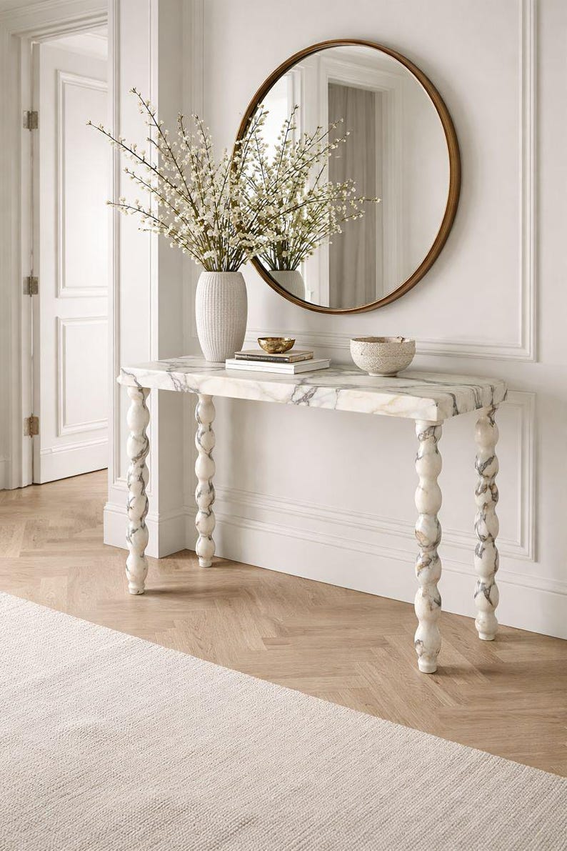 May include: A marble-topped console table with turned legs, a round mirror, and a vase of white flowers. The table has a rectangular top with grey veining and decorative, white and grey legs. A gold bowl and a textured bowl sit on the table.