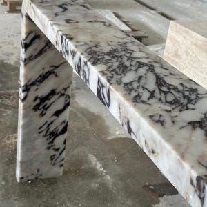 May include: A waterfall console table crafted from white marble with prominent, dark purple and black veining. The piece features a smooth, polished surface and a continuous, seamless L-shaped design that connects the top to a single side leg.