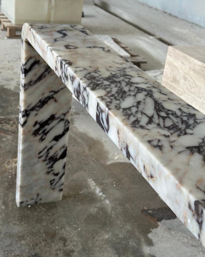 May include: A waterfall console table crafted from white marble with prominent, dark purple and black veining. The piece features a smooth, polished surface and a continuous, seamless L-shaped design that connects the top to a single side leg.