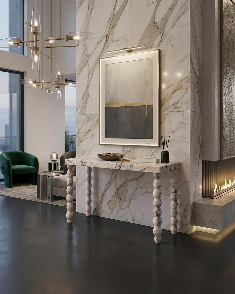 May include: A contemporary living room featuring a marble console table with turned legs, an abstract framed artwork, and a green armchair. The table holds a bowl and a black vase. A fireplace is also present.