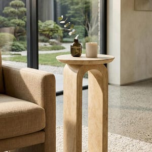 May include: A light-colored, arched side table with a round top. A small brown glass bottle with greenery and a white candle sit on the table. The table is next to a beige armchair.
