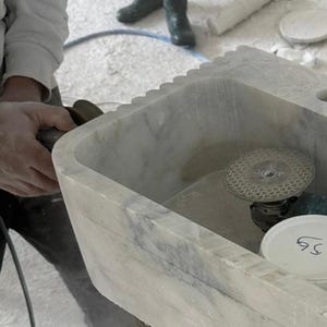 Puede incluir: Un lavabo de m&aacute;rmol gris claro con borde festoneado est&aacute; siendo trabajado con una herramienta el&eacute;ctrica. El lavabo tiene una cubeta rectangular y est&aacute; en proceso de elaboraci&oacute;n. Una persona sostiene la herramienta el&eacute;ctrica.