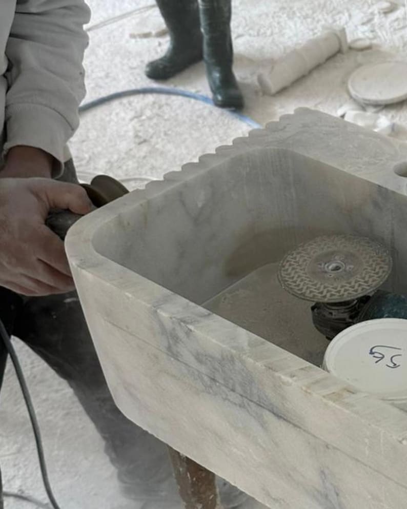 Puede incluir: Un lavabo de m&aacute;rmol gris claro con borde festoneado est&aacute; siendo trabajado con una herramienta el&eacute;ctrica. El lavabo tiene una cubeta rectangular y est&aacute; en proceso de elaboraci&oacute;n. Una persona sostiene la herramienta el&eacute;ctrica.