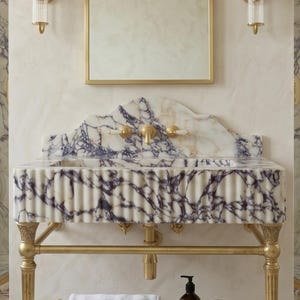 Pu&ograve; includere: Un mobile lavabo a parete con una vasca in marmo bianco venato di viola e frontale scanalato, sostenuto da una struttura in ottone. Il set include un paraschizzi in marmo abbinato, un rubinetto in ottone e due applique da parete.