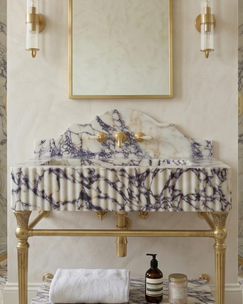 Pu&ograve; includere: Un mobile lavabo a parete con una vasca in marmo bianco venato di viola e frontale scanalato, sostenuto da una struttura in ottone. Il set include un paraschizzi in marmo abbinato, un rubinetto in ottone e due applique da parete.