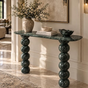 Op de afbeelding: Een donkergroene marmeren sidetable met een glad, langwerpig ovaal blad, ondersteund door twee verticale kolommen bestaande uit gestapelde marmeren bollen. Op het tafelblad staan een textuurrijke vaas met groen, twee boeken en een schaal.
