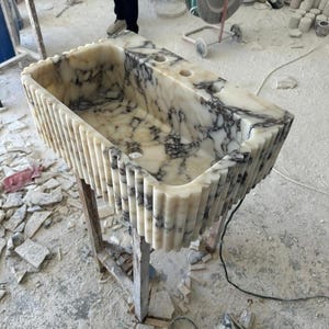 Puede incluir: Un lavabo rectangular de m&aacute;rmol con exterior estriado. El lavabo es blanco con vetas grises y tiene dos orificios para grifos. Est&aacute; colocado sobre un soporte de madera en un taller. Las dimensiones del lavabo no son visibles.