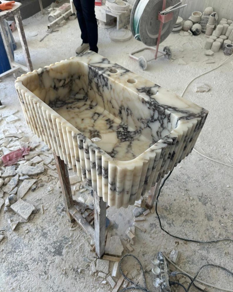 Puede incluir: Un lavabo rectangular de m&aacute;rmol con exterior estriado. El lavabo es blanco con vetas grises y tiene dos orificios para grifos. Est&aacute; colocado sobre un soporte de madera en un taller. Las dimensiones del lavabo no son visibles.