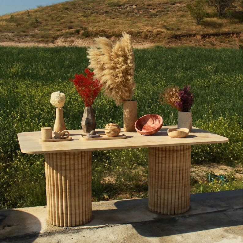 Puede incluir: Una mesa de madera clara con dos patas cil&iacute;ndricas. La mesa est&aacute; decorada con jarrones con flores secas, un cuenco y peque&ntilde;os objetos de madera. El fondo presenta un campo verde y un cielo azul.