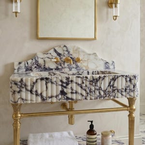 Pu&ograve; includere: Un lavabo in marmo bianco e viola con frontale scanalato, sostenuto da gambe in metallo dorato. Il lavabo presenta un paraschizzi e un ripiano in marmo abbinati, un rubinetto dorato e due applique dorate sopra uno specchio rettangolare.