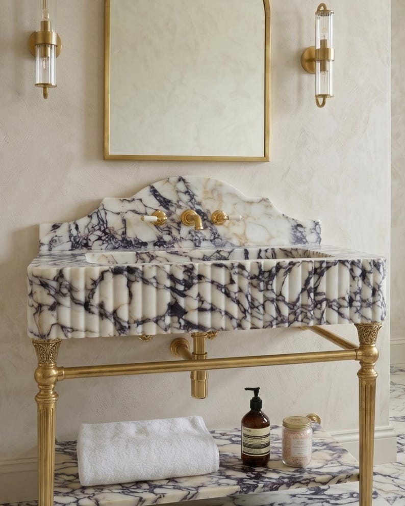 Pu&ograve; includere: Un lavabo in marmo bianco e viola con frontale scanalato, sostenuto da gambe in metallo dorato. Il lavabo presenta un paraschizzi e un ripiano in marmo abbinati, un rubinetto dorato e due applique dorate sopra uno specchio rettangolare.