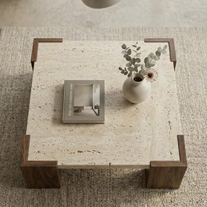 May include: A square coffee table with a beige porous travertine stone top and dark brown wooden corner legs. A small beige ceramic vase containing dried eucalyptus stems and a book titled Wabi-Sabi architecture notes rest on the surface.