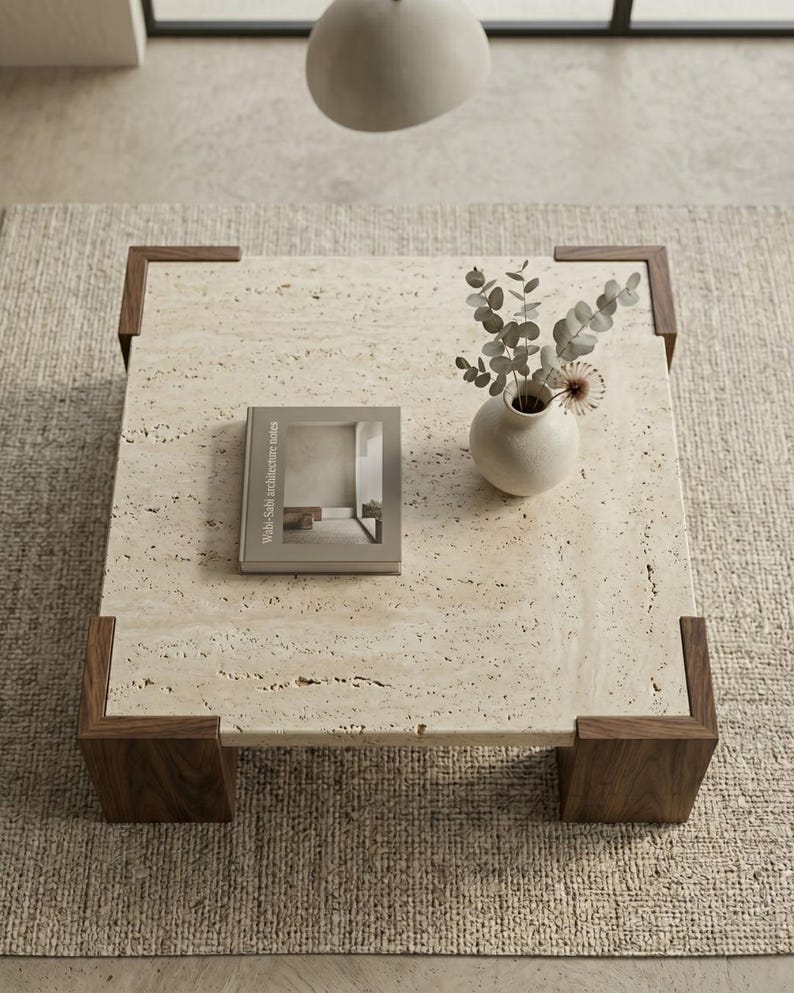 May include: A square coffee table with a beige porous travertine stone top and dark brown wooden corner legs. A small beige ceramic vase containing dried eucalyptus stems and a book titled Wabi-Sabi architecture notes rest on the surface.