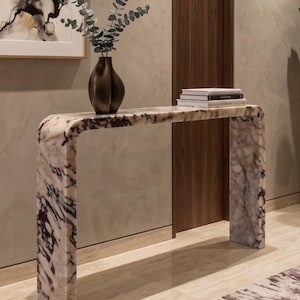 May include: A waterfall console table crafted from white marble with prominent dark purple and grey veining. A dark brown sculptural vase containing eucalyptus branches and a small stack of white books sit on the smooth, polished surface.