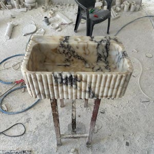 Puede incluir: Un lavabo rectangular de m&aacute;rmol con exterior estriado e interior veteado negro y beige. El lavabo est&aacute; sostenido por un soporte met&aacute;lico. El fondo muestra una silla de pl&aacute;stico negro y diversos materiales de construcci&oacute;n.