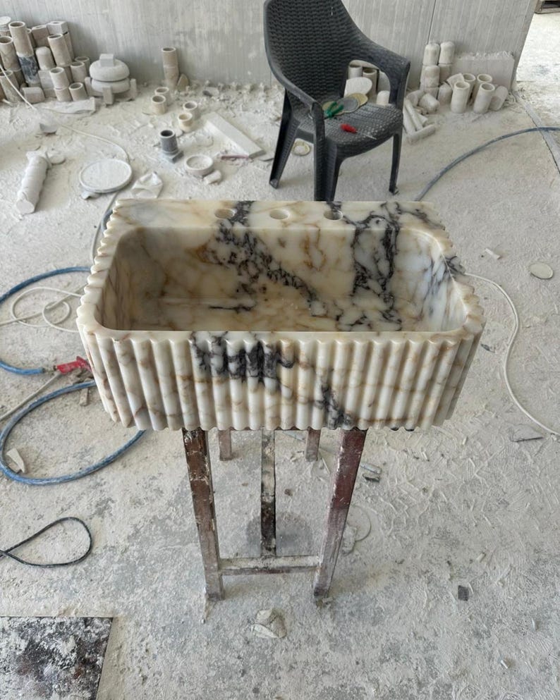 Puede incluir: Un lavabo rectangular de m&aacute;rmol con exterior estriado e interior veteado negro y beige. El lavabo est&aacute; sostenido por un soporte met&aacute;lico. El fondo muestra una silla de pl&aacute;stico negro y diversos materiales de construcci&oacute;n.