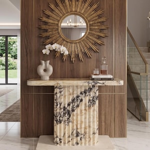 Pode incluir: Um espelho dourado em forma de sol está pendurado sobre uma mesa de consola com uma base canelada de mármore creme e preto e um tampo liso. Na mesa, há um vaso de cerâmica branca com orquídeas, um decantador, um copo e dois livros.
