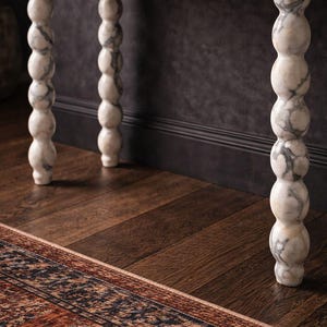 May include: A marble-topped console table with four decorative, turned legs. The legs are white with grey veining. The table sits on a dark wood floor, and a patterned rug is visible in the foreground. The wall behind the table is dark grey.