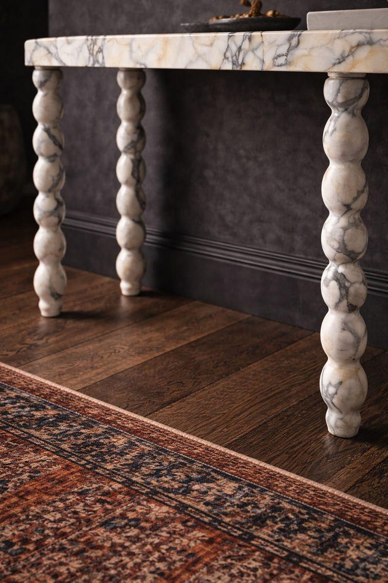 May include: A marble-topped console table with four decorative, turned legs. The legs are white with grey veining. The table sits on a dark wood floor, and a patterned rug is visible in the foreground. The wall behind the table is dark grey.