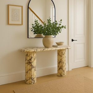 May include: A marble console table with fluted legs, topped with a vase of greenery, a small bowl, and a black arched mirror. A framed abstract art piece hangs on the wall. A tan rug is on the floor.