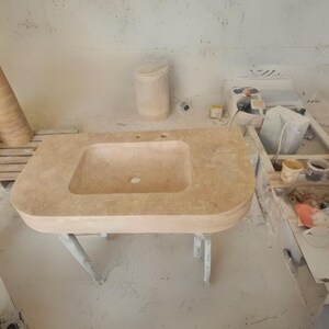 May include: A light beige rectangular stone sink with a built-in basin and two tap holes. The sink is supported by a white metal frame with four legs. The background shows a workshop setting.