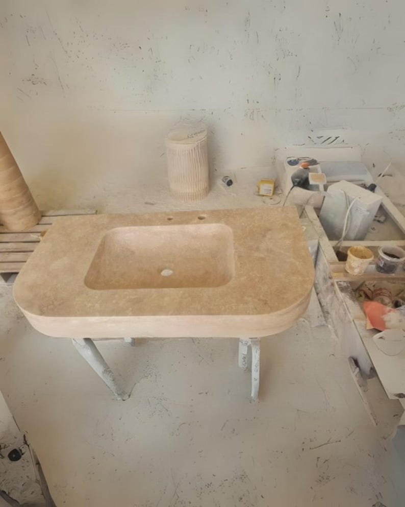 May include: A light beige rectangular stone sink with a built-in basin and two tap holes. The sink is supported by a white metal frame with four legs. The background shows a workshop setting.