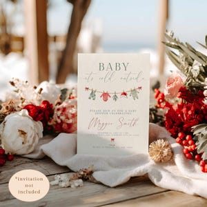 May include: A baby shower invitation with the text "BABY it's cold outside." The invitation features a clothesline with baby clothes illustrations. The invitation is decorated with flowers and greenery.