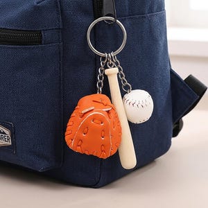 Könnte beinhalten: Ein marineblauer Rucksack mit schwarzem Reißverschluss und einem silbernen Schlüsselring. Am Schlüsselring hängen Baseball-Anhänger: ein orangefarbener Baseballhandschuh, ein Baseballschläger aus Holz und ein weißer Baseball.